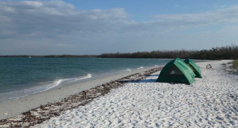 Two tents rest on a sandy beach beside a blue body of water. 